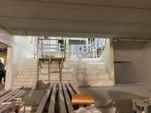 Een nog onaffe ruimte met daarin bouwmaterialen en een steiger. Brede trappen leiden naar de hoger gelegen verdieping.