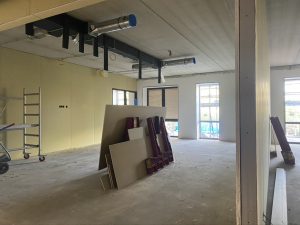 Een lege, nog niet afgebouwde ruimte met in het midden platen bouwmateriaal. De wanden zijn geel en in de linkerhoek staat een lage steiger.