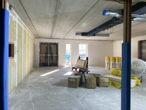 Een grote lege ruimte in afbouw met daarin twee ramen van glas en nog afgedekte openslaande deuren. Er staan gele rollen isolatiemateriaal en dozen.