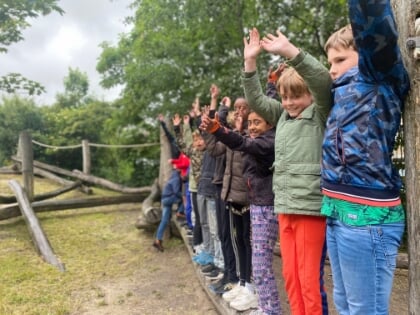 Op een boomstam in een natuurspeeltuin staan acht kinderen op een rij. Ze doen de wave. Op de achtergrond zijn bomen zichtbaar en meer natuurlijke speelmaterialen.