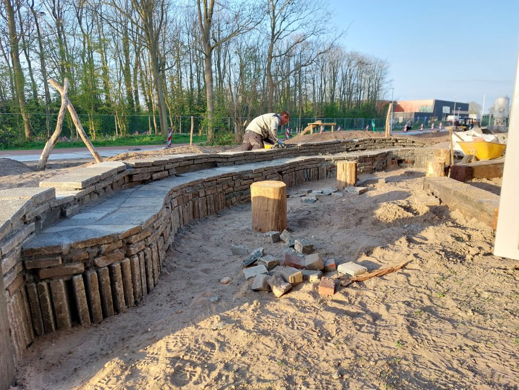 Op de voorgrond een zandondergrond waarop een muurtje wordt gemetseld dat in een golvende vorm loopt. Het lijkt op een lange bank en wordt straks een buitenklas. Op de achtergrond bomen en een speelelement van hout.