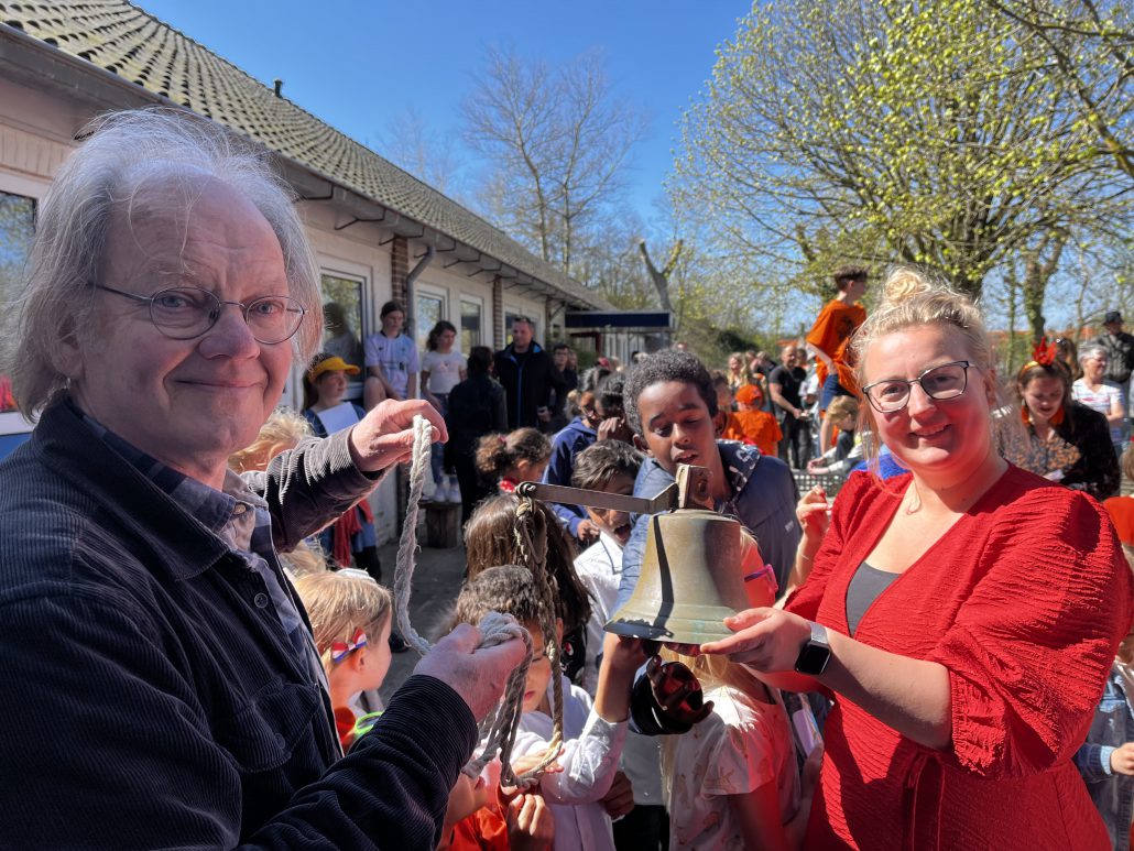Een blonde vrouw in een rode jurk draagt een schoolbel over aan een oudere heer die op de voorgrond lachend in de camera kijkt. Op de achtergrond staan schoolkinderen en het schoolgebouw.