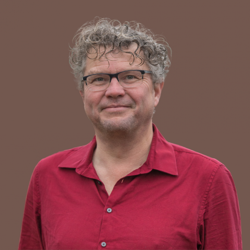 Wethouder Remko van de Belt kijkt lachend de camera in. Hij draagt een rood overhemd. De achtergrond is donker.
